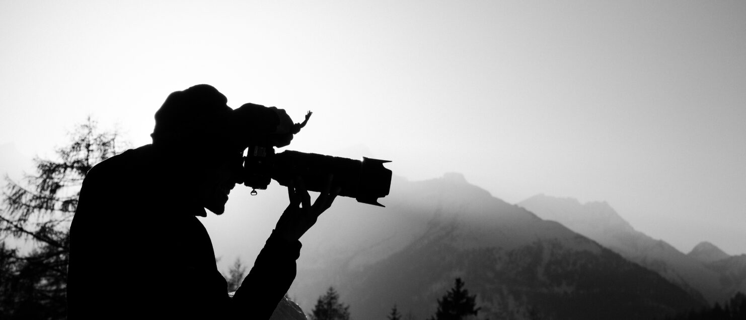 Schwarz-Weiß-Silhouette eines Fotografen mit Teleobjektiv vor Bergkulisse – klassische Bergfotografie
