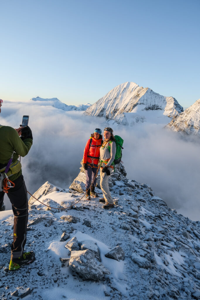 Zwei Bergsteiger posieren am Gipfelgrat des Ortler über dem Nebelmeer, eine Person fotografiert mit dem Smartphone